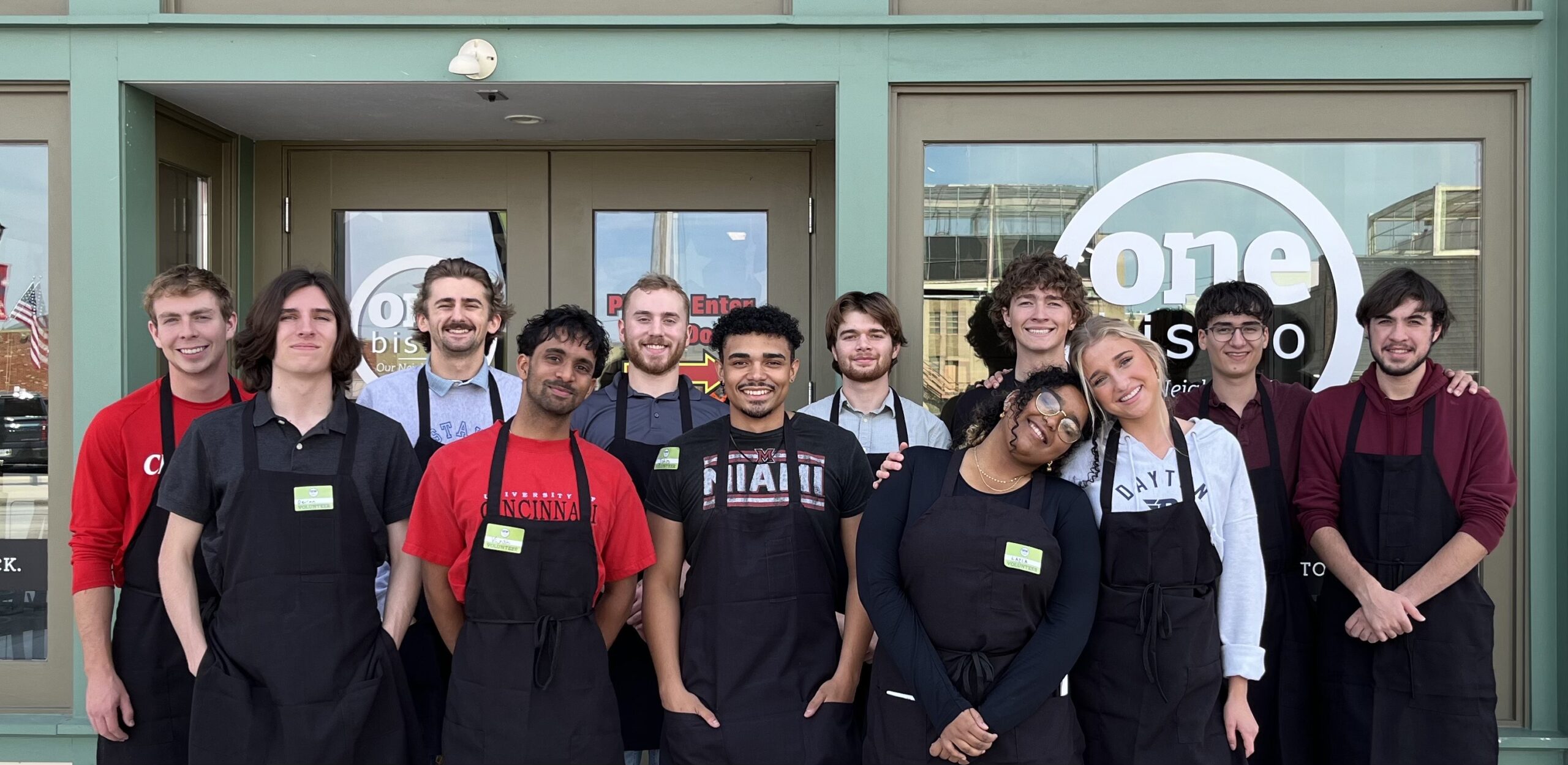 A group of thirteen people wearing black aprons stand smiling in front of a restaurant with glass doors and a sign that says one bistro..