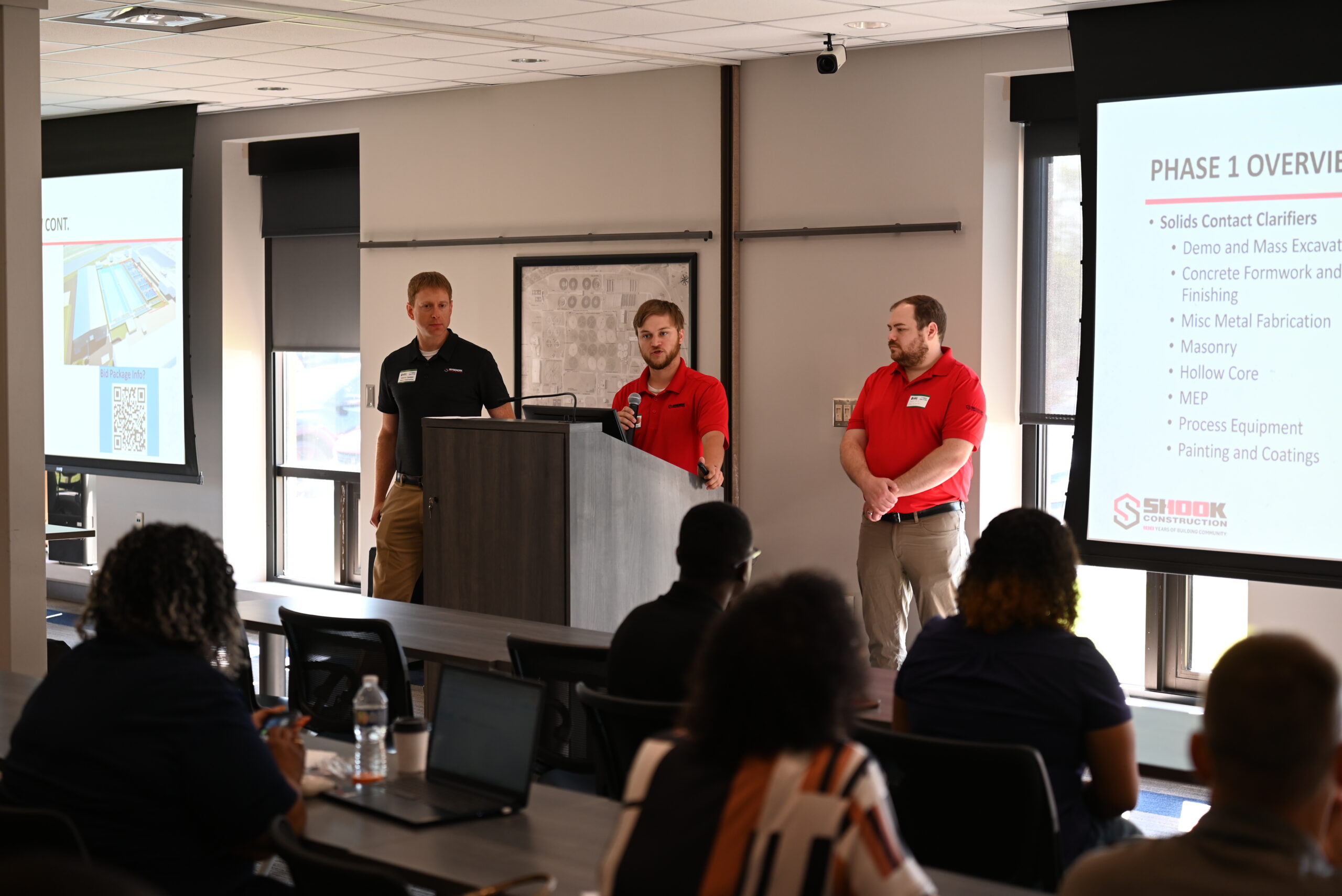 Three men in red and black shirts present at a podium in a classroom, while an audience listens. Presentation slides titled Phase 1 Overview display project details on screens at the front of the room.