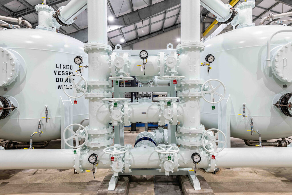 Industrial piping system with multiple white pipes, pressure gauges, and valves inside a large facility. Two large cylindrical tanks with labels and a metal roof are visible in the background.