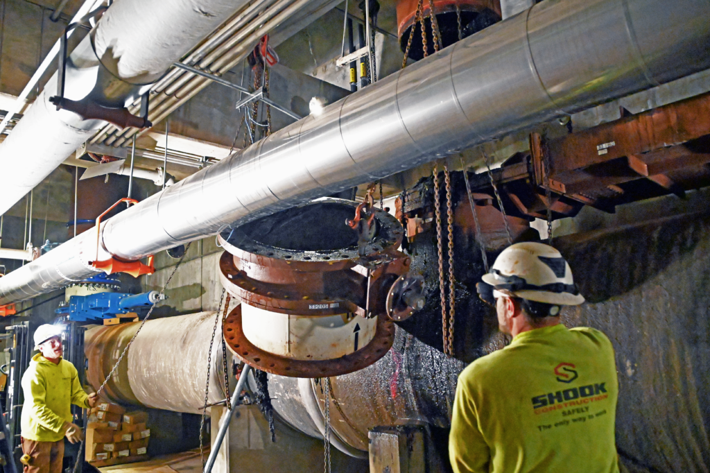 Two workers in safety gear install a large industrial pipe section in an underground facility. One worker observes as the pipe is being positioned with a chain hoist, while the other stands nearby.