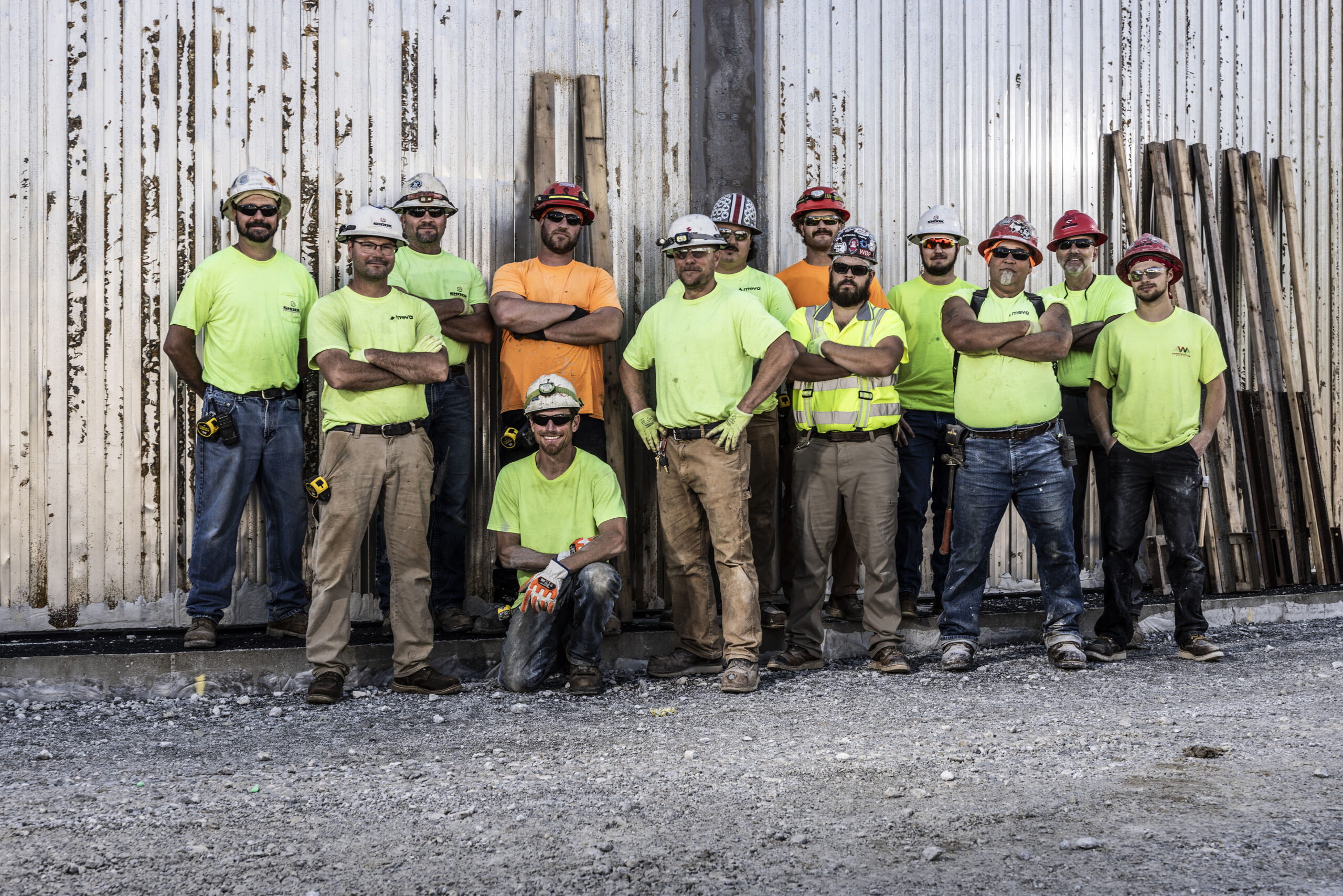 Twelve construction workers in safety gear and neon shirts stand and kneel in front of a corrugated metal wall with wooden planks, posing for a group photo outdoors at a worksite.