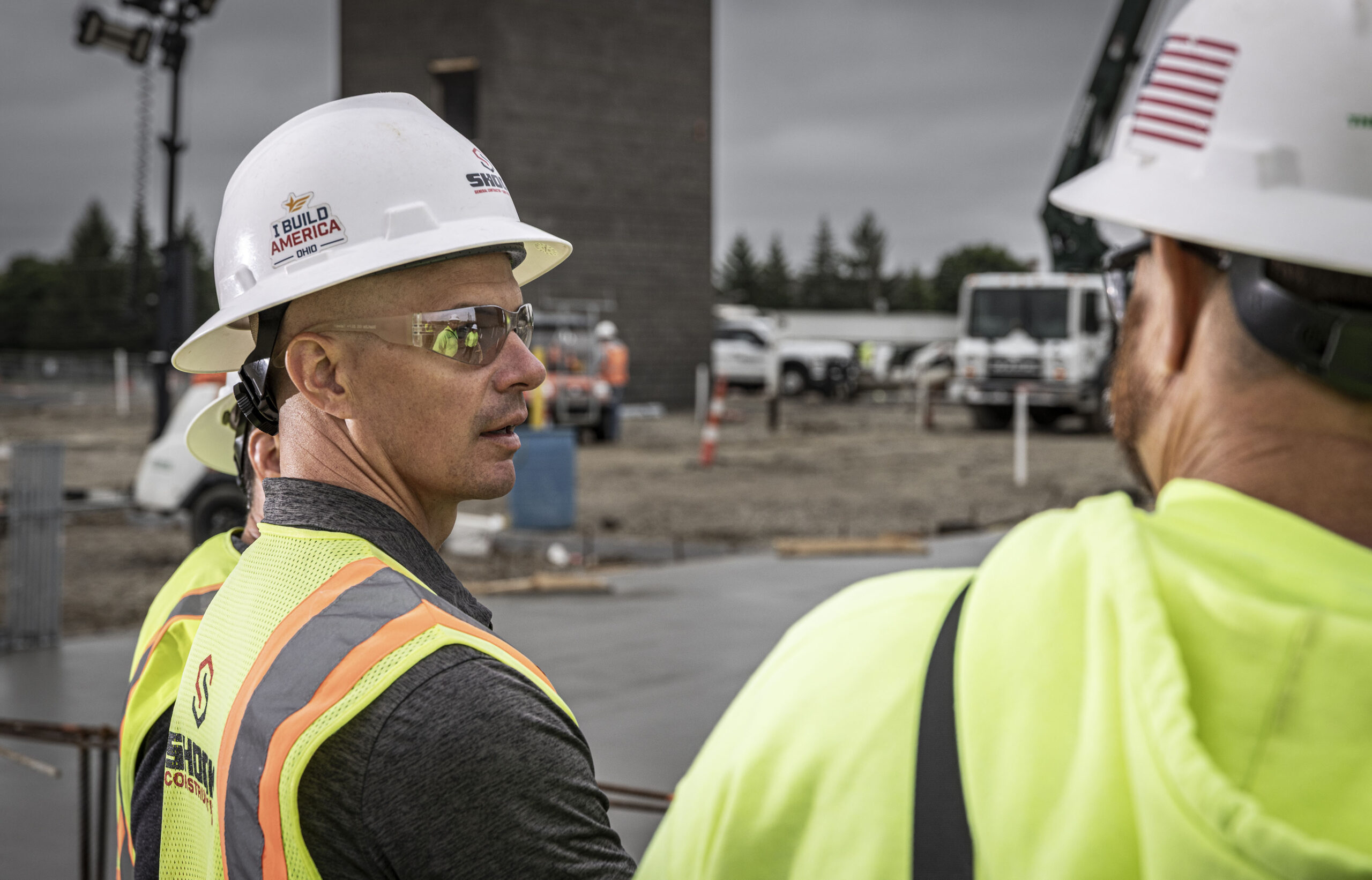Two construction workers wearing hard hats and safety vests talk at an outdoor job site. One worker faces the camera; the other is seen from behind. Construction equipment and vehicles are visible in the background.