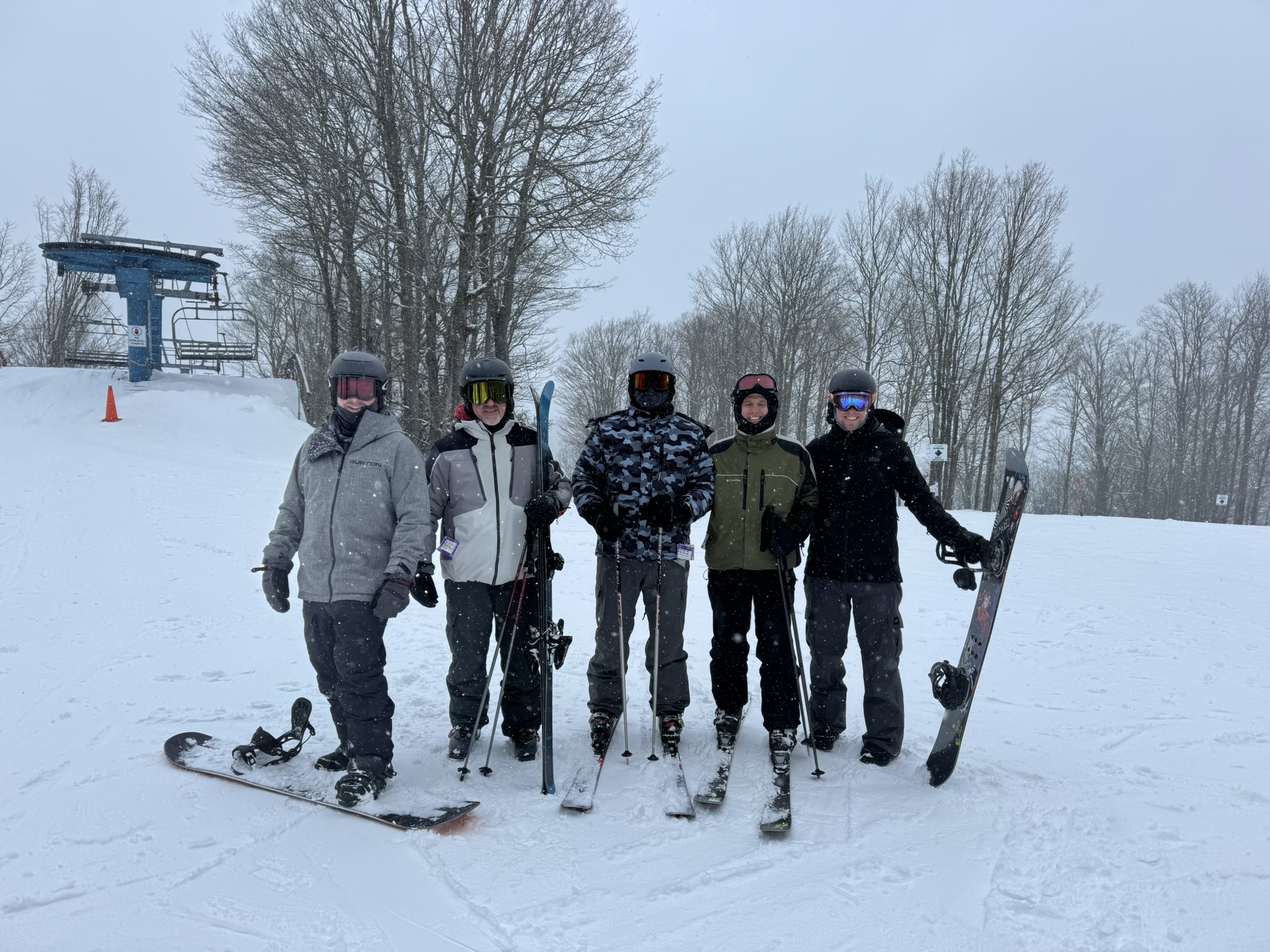 Five people in winter gear stand on a snowy ski slope with skis and snowboards. Bare trees and a ski lift are in the background on a cloudy day.