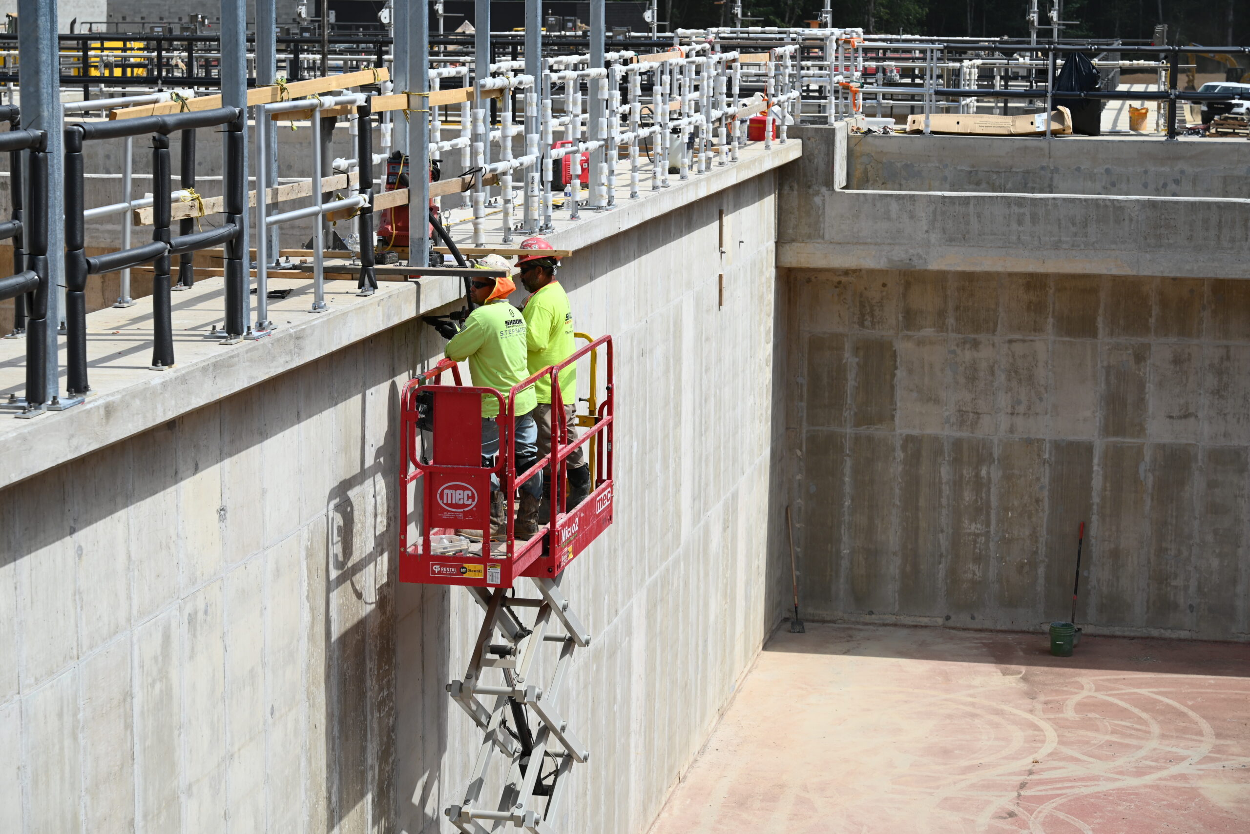 Two construction workers in neon shirts use a red scissor lift to work on pipes mounted to a tall concrete wall at an industrial site. The ground below is bare concrete with a broom and green container.
