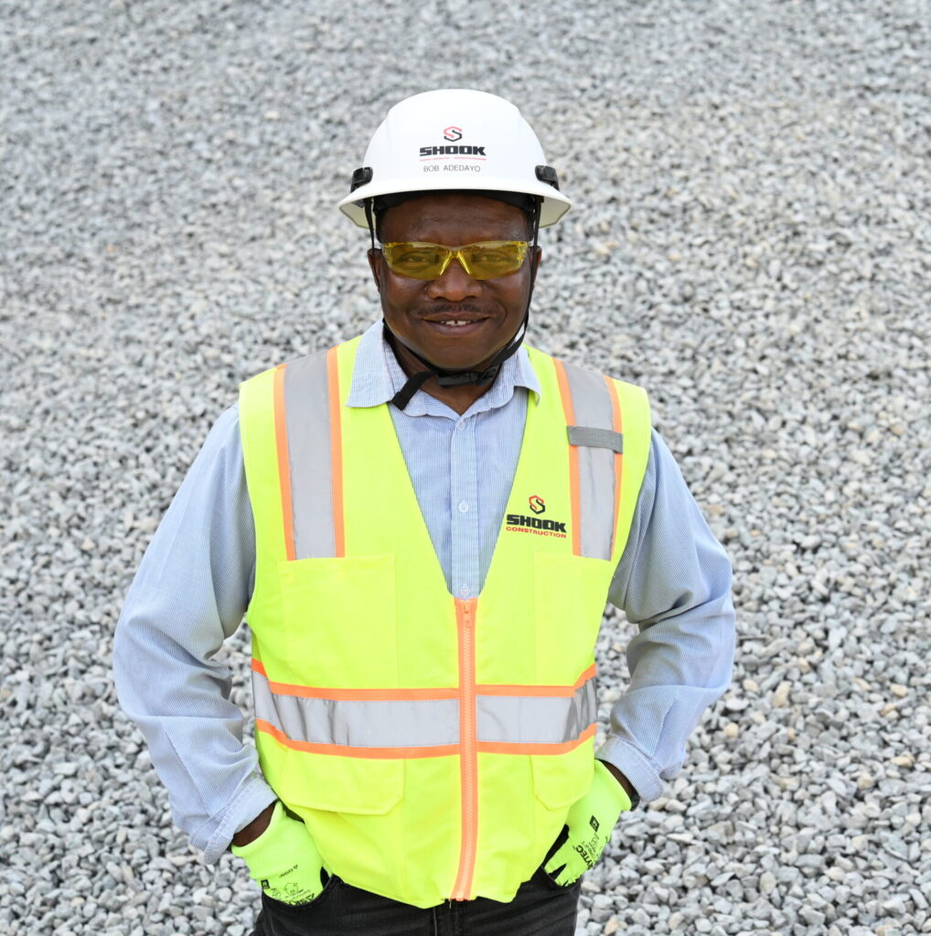 A construction worker wearing a white hard hat, yellow safety vest, gloves, and yellow safety glasses stands smiling on a gravel surface with hands in pockets.