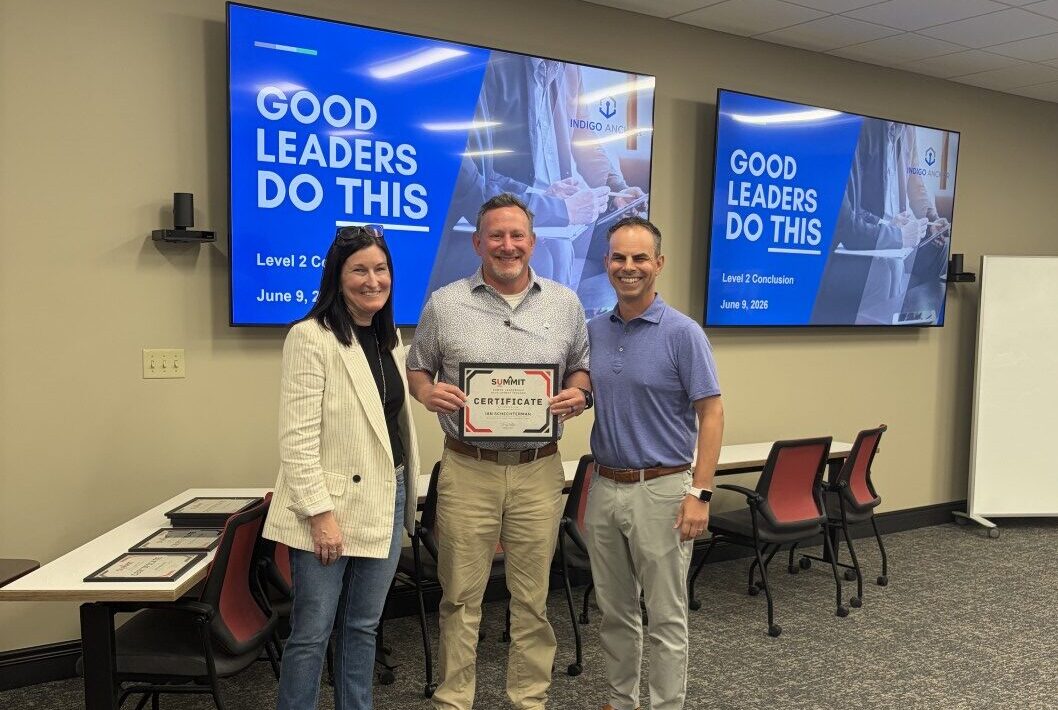Three people stand in a classroom. The person in the middle holds a certificate. Behind them, two screens display Good Leaders Do This and course details.