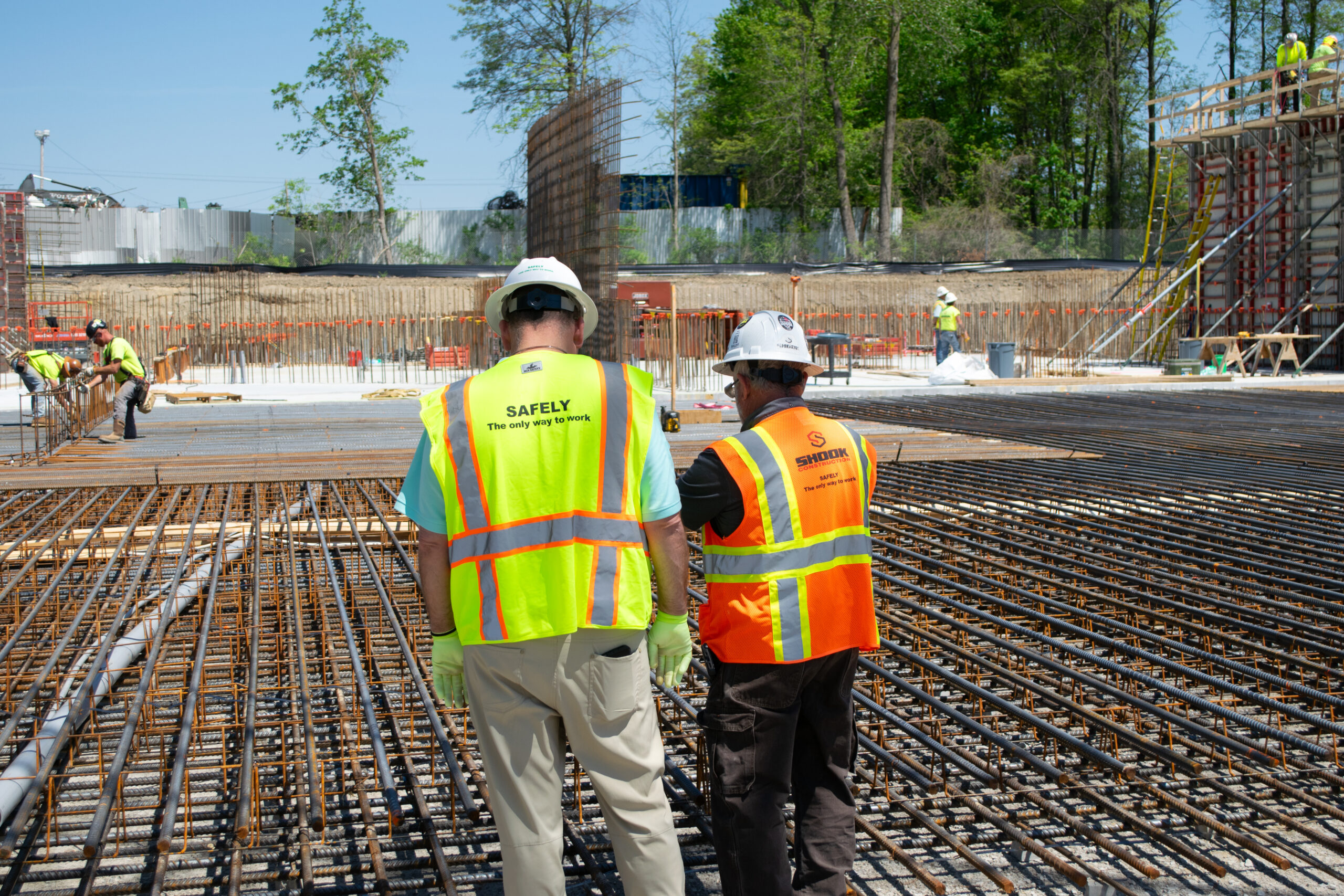 Two construction workers in safety vests and helmets stand on a site with exposed rebar, overseeing progress as others work in the background; trees and concrete walls are visible.