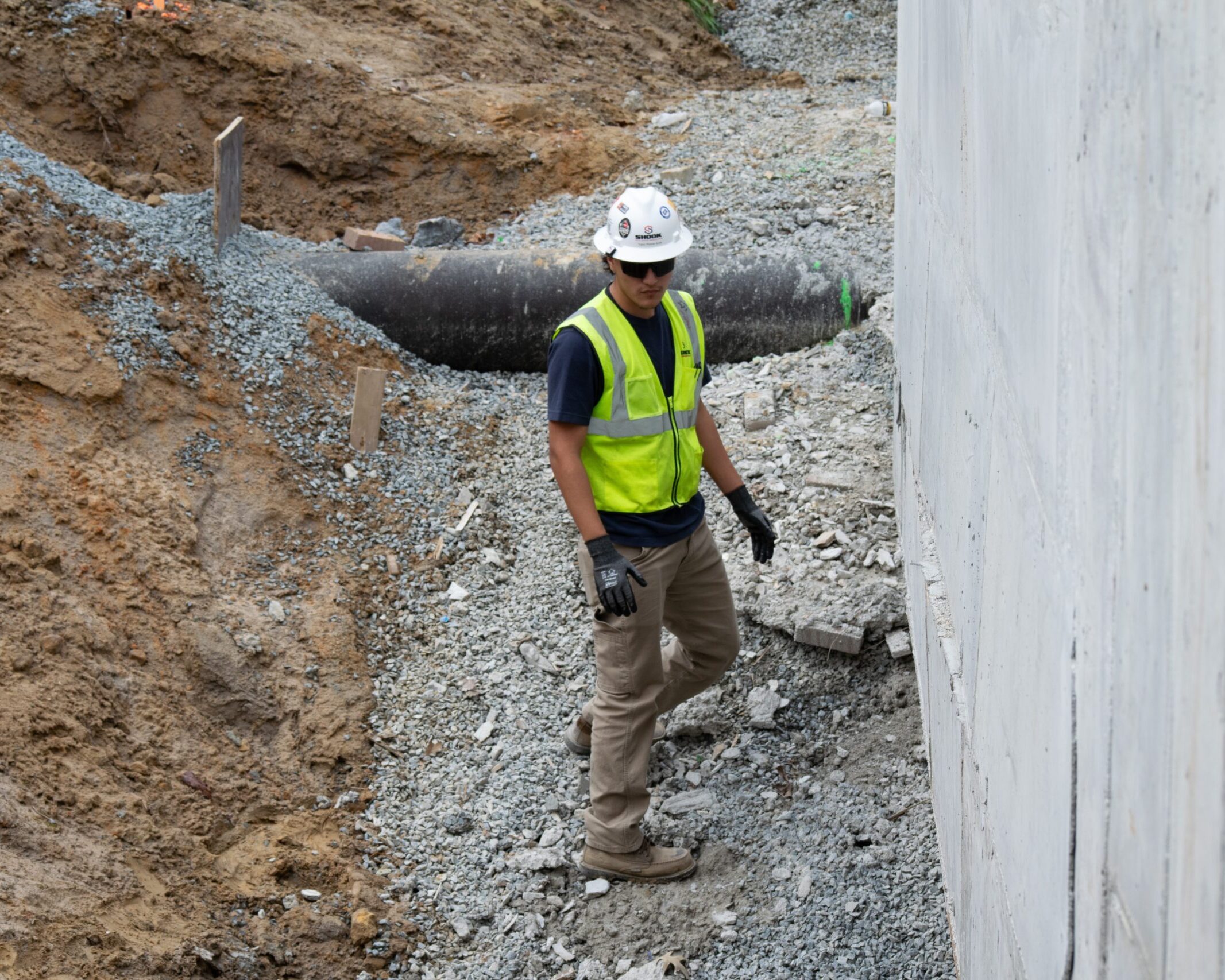 A construction worker in a hard hat, safety vest, and gloves walks beside a large concrete wall at a gravel-covered construction site with exposed pipes and dirt.